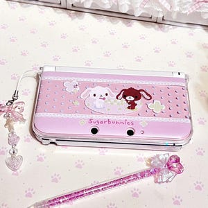 May include: A pink Nintendo 3DS console with a Sugarbunnies design, featuring two cartoon bunnies, clouds, and lace details. A matching pen with pink glitter and a charm are also present. The background is a paw print pattern.