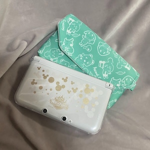 Può includere: Una console di gioco portatile Nintendo 3DS bianca con disegni dorati di Mickey Mouse e castello. Una custodia verde menta con personaggi di cartoni animati bianchi è accanto alla console. La console è su una superficie grigia.