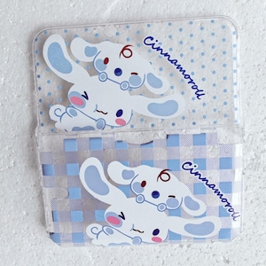 May include: Clear plastic card holder featuring a white cartoon dog with blue accents and the word "Cinnamoroll" in blue script. The background has a blue and white polka dot and plaid pattern.