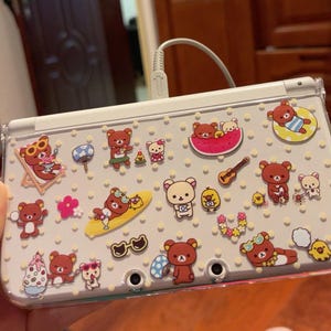 Puede incluir: Una consola de juegos portátil blanca con una funda protectora transparente. La funda está decorada con numerosas pegatinas de osos de dibujos animados en varias poses, junto con otros gráficos con temática veraniega. Las pegatinas son en tonos marrón, amarillo y rosa.