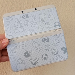 Puede incluir: Consola de juegos portátil Nintendo 3DS de color gris claro, abierta para revelar la pantalla. La consola está decorada con un patrón de personajes de dibujos animados blancos e iconos de la serie de videojuegos Super Mario Bros.