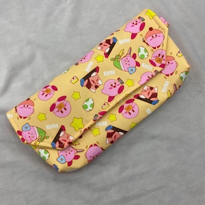 Puede incluir: Un estuche para gafas rectangular amarillo con un patrón repetido de personajes rosas de Kirby, estrellas y huevos. El estuche tiene un cierre de solapa y la palabra "Kirby" está impresa.