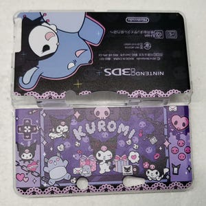 May include: A clear plastic Nintendo 3DS case featuring a purple and black design with the character Kuromi. The case has a decorative border and includes the text "KUROMI" and "Nintendo 3DS+".