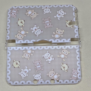 Op de afbeelding: Een doorzichtige plastic Nintendo 3DS-hoes met een beige achtergrond met herhaalde illustraties van cartoonberen en kuikens. De hoes heeft een witte, kantachtige rand met hartvormige uitsparingen.