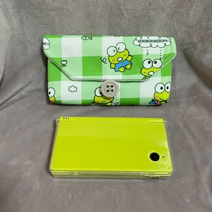 以下が含まれることがあります： 透明な保護ケースに入ったライムグリーンのニンテンドーDS Liteコンソールが、緑と白のケロピをテーマにしたケースの下にあります。ケースにはボタン留めと、ケロピのキャラクターと「KEROKEROKEROPPI」の文字が描かれています。