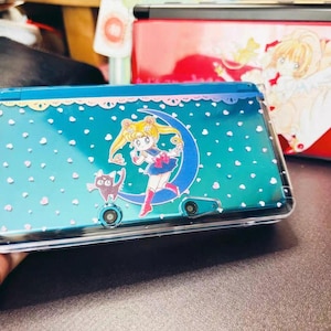 Puede incluir: Una consola Nintendo 3DS turquesa con una funda protectora transparente. La consola presenta un diseño de Sailor Moon con una luna creciente, un gato y detalles en forma de corazón. En el fondo se ve otra consola roja con un diseño blanco.