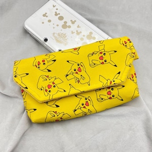 Puede incluir: Clutch amarilla con un patrón de Pikachu, con contornos negros y detalles rojos. Una Nintendo 3DS blanca con diseños de Mickey Mouse es parcialmente visible en el fondo. La clutch está doblada.