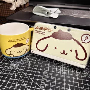 Puede incluir: Se muestra una taza de cerámica amarilla y un dispositivo de juego portátil de color crema. La taza presenta un diseño de perro de dibujos animados y el texto "Every day's a fun day together with my friends." El dispositivo tiene un diseño de perro de dibujos animados.