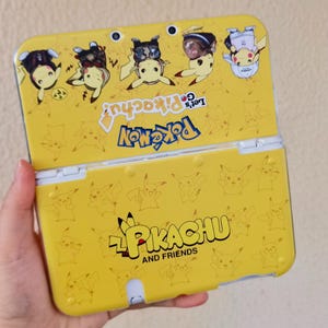 Puede incluir: Funda amarilla para Nintendo 3DS con Pikachu y otros personajes de Pokémon. La funda tiene el texto "Let's Go Pikachu!" y "Pikachu and Friends". La funda es amarilla con bisagras blancas.