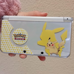 Puede incluir: Consola de juegos portátil Nintendo 3DS blanca con diseño de Pikachu. La consola presenta una silueta amarilla de Pikachu y un gráfico de Pikachu más pequeño. También es visible el logotipo de Pokémon Trading Card Game.