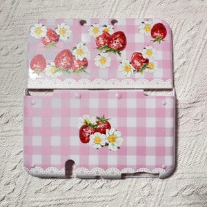 Può includere: Una custodia rosa e bianca a quadretti per Nintendo 3DS con un design a fragole e fiori. La custodia è decorata con fragole rosse, margherite bianche e un bordo in pizzo bianco. La custodia è aperta.