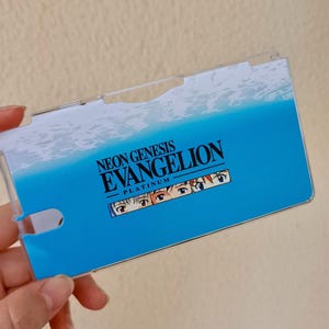 Puede incluir: Una funda de plástico transparente con un diseño degradado azul y blanco. La funda presenta el texto "NEON GENESIS EVANGELION PLATINUM" en negro y una fila de ojos de personajes. La funda está sujeta por una mano.