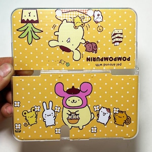 Puede incluir: Funda amarilla con lunares blancos con personajes de dibujos animados. La funda tiene un marco de plástico transparente y un diseño con bisagras. Los personajes incluyen un oso con un sombrero rosa, un conejo y un gato. El texto "POMPOMPURIN" es visible.