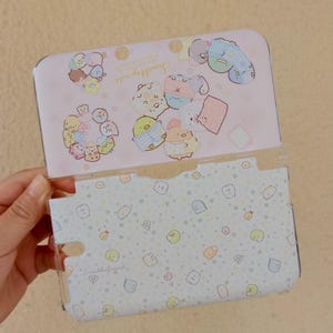 May include: A clear plastic case for a handheld gaming device, featuring a light pink and white design with cartoon characters. The case has a hinged top and a patterned interior with small, colorful illustrations.