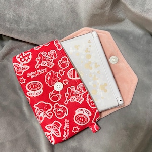 Może przedstawiać: Czerwone i białe etui na Nintendo DS Super Mario Move cat z różowym wnętrzem. Etui posiada magnetyczne zapięcie i białą konsolę Nintendo DS w środku. Etui leży na szarej powierzchni.