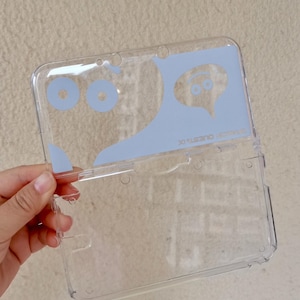 Puede incluir: Una funda protectora de plástico transparente para consola de juegos portátil. La sección superior presenta un estampado gráfico azul claro de dos ojos circulares y un globo de texto con una cara sonriente, con el texto DRAGON QUEST IX.