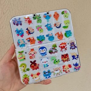 May include: A clear protective case for a Nintendo 3DS LL handheld game console, adorned with colorful cartoon character stickers. The stickers feature various creatures in shades of blue, orange, green, and red. The console is held open, displaying the screen.