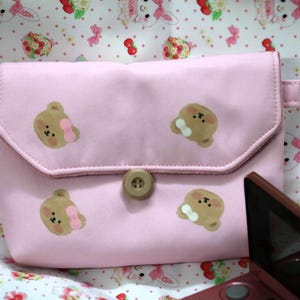 May include: A pink, rectangular pouch with a flap closure and a button. The pouch is decorated with brown bear illustrations, some with pink bows. The background features a patterned fabric with bunnies and cherries.