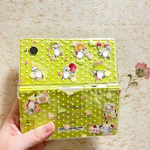 Puede incluir: Una consola de juegos portátil Nintendo DS Lite verde lima con una cubierta transparente adornada con pegatinas de personajes de dibujos animados e ilustraciones de frutas. Las pegatinas incluyen personajes con sombreros de frutas y alimentos. La consola se sostiene en una mano.