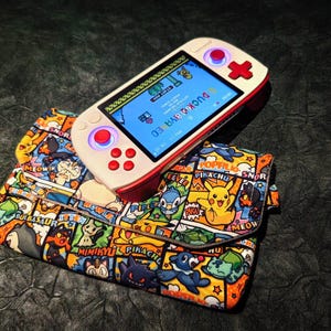 May include: A white and red handheld gaming device with a screen displaying a game interface rests on a colourful wallet. The wallet features a comic book style design with various cartoon characters in vibrant colours. The device has red buttons and a directional pad.