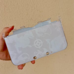 May include: A clear protective case for a handheld gaming device. The light grey case features a repeating dragon pattern and a central emblem. It has cutouts for buttons and ports. The case is held in a hand.