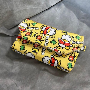 Puede incluir: Un bolso de mano amarillo con un diseño de pato de dibujos animados. El bolso tiene un cierre de solapa y está decorado con imágenes de un pato blanco con una camisa roja, con la palabra "Pekkje".