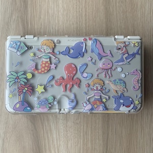 May include: A light grey Nintendo 3DS case decorated with cartoon illustrations of mermaids, whales, an octopus, a seahorse, a turtle, and a shark. The case has a clear plastic overlay.