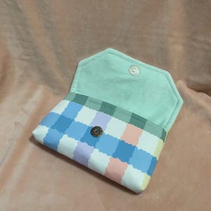 May include: Open pastel-coloured wallet with a gingham pattern in blue, pink, and yellow. The interior is a light green fabric. The wallet is secured with a silver snap closure.