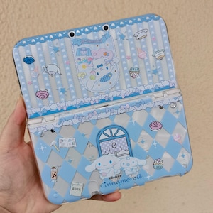 Op de afbeelding: Een lichtblauwe Nintendo 3DS-hoes met een Cinnamoroll-ontwerp. De hoes heeft een herhalend patroon van ruiten en strikken, met illustraties van desserts en andere schattige elementen. De tekst "Cinnamoroll" is onderaan zichtbaar.