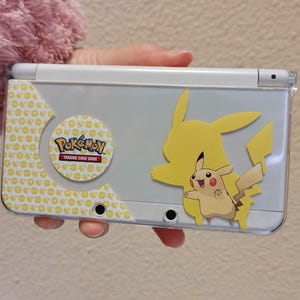 Puede incluir: Consola de juegos portátil gris con diseño de Pikachu. La consola tiene detalles amarillos y una silueta de Pikachu. También es visible una pegatina circular con el logo "Pokémon Trading Card Game".