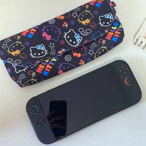May include: A black clutch bag and a black games console. The clutch has a repeating pattern of Hello Kitty faces, stars, and the words "Hello Kitty" in rainbow colours. The games console is black with grey and orange accents.