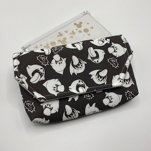 Può includere: Una pochette a fantasia bianca e nera con chiusura a patta, con un motivo ripetuto di fantasmi dei cartoni animati. Un dispositivo di gioco portatile bianco con silhouette dorate di Topolino è parzialmente visibile sotto.