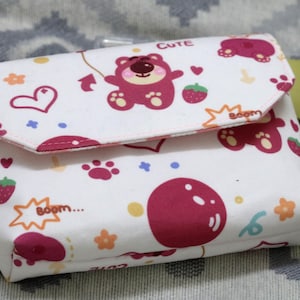 May include: A white fabric wallet with a flap closure, decorated with cartoon bears, strawberries, hearts, and the word "CUTE". The design is in shades of pink and red. The wallet has a snap closure.