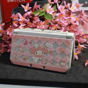 Puede incluir: Una funda rosa y blanca para Nintendo 3DS con un diseño de My Melody. La funda tiene un patrón de cuadros con lazos, corazones y la palabra "Cute". La funda está decorada con detalles florales rosas y blancos.