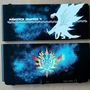 Può includere: Due custodie nere per cartucce di gioco Nintendo 3DS. Una presenta il testo "MONSTER HUNTER 4" con una silhouette di mostro bianca. L'altra custodia ha un design con emblema colorato su sfondo blu.