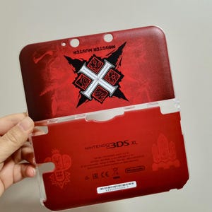 Puede incluir: Funda roja para Nintendo 3DS XL con diseño de Monster Hunter. La cubierta superior presenta un emblema en blanco y negro sobre un fondo rojo texturizado. La cubierta inferior también es roja con el logotipo de Nintendo 3DS XL.