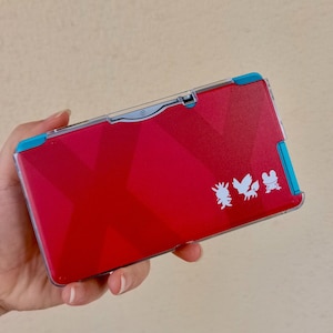 Puede incluir: Una consola de juegos portátil roja con un diseño geométrico y siluetas de personajes blancos. La consola tiene detalles en azul y una funda protectora transparente. El dispositivo se sostiene en una mano.