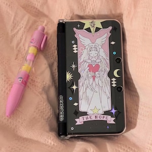 May include: A black Nintendo 3DS handheld gaming console with a clear protective case featuring a colorful illustration of an angel with wings and a heart. The text "THE HOPE" is printed on the case.
