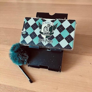 Könnte beinhalten: Eine schwarze Nintendo DS-Hülle mit dem Slytherin-Wappen und dem Schriftzug "Harry Potter". Die Hülle hat ein rautenförmiges Muster in Schwarz und Türkis. Ein grüner, flauschiger Stift liegt daneben.