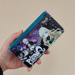 Könnte beinhalten: Ein teal Nintendo 3DS Handheld-Spielkonsole mit einer individuellen Hülle mit Anime-Charakteren und dem Text "Persona Q Shadow of the Labyrinth". Das Cover-Design enthält ein Schachbrettmuster und verschiedene Charakterillustrationen.