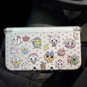 May include: A white Nintendo 3DS case with a clear plastic cover, adorned with colorful cartoon character stickers and pink polka dots. The case has three circular holes at the bottom and is closed.