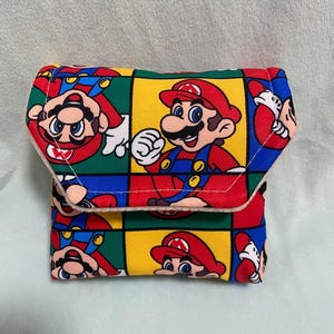 May include: A small, square wallet with a colourful pattern of the video game character Mario. The design includes multiple images of Mario in red, blue, yellow, and green squares. The wallet has a fold-over flap.