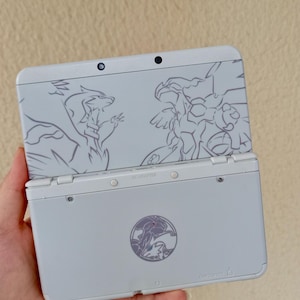 Puede incluir: Consola de juegos portátil Nintendo 3DS de color gris claro. La cubierta superior presenta un contorno detallado de dos criaturas míticas. La parte inferior de la consola tiene un emblema circular y el texto "GAME CARD".