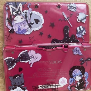 May include: A pink Nintendo 3DS handheld gaming console with a clear plastic case. The case is decorated with black and white cartoon characters, hearts, stars, and the words "Hate" and "Happy".