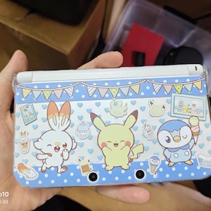Może przedstawiać: Niebiesko-białe etui na Nintendo 3DS z postaciami z kreskówek. Etui ma jasnoniebieskie tło z białymi kropkami i wzorem banera. Postacie to królik, Pikachu i pingwin. Etui jest trzymane w dłoni.