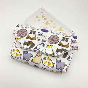 May include: A white clutch with a repeating pattern of cartoon cats in various poses and colors. The clutch is folded over and secured with a flap. A clear case with gold Mickey Mouse designs is behind the clutch.