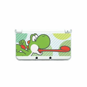May include: White Nintendo 3DS handheld game console featuring a green Yoshi graphic. The design shows Yoshi with his tongue extended, set against a background of green and blue circles and stripes.
