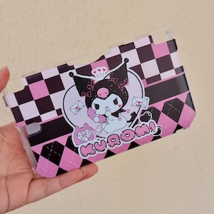 May include: A clear phone case with a pink, black, and white chequered design. The case features a cartoon character graphic with a heart-shaped face, holding a phone, and the word "Kuromi" in pink.