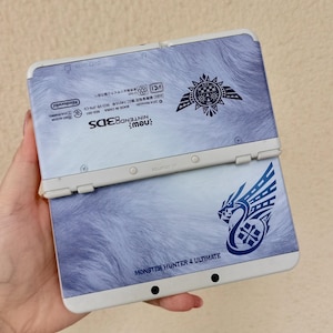 May include: A New Nintendo 3DS handheld gaming console featuring a light blue and white fur-like pattern. The device displays the Monster Hunter 4 Ultimate logo, a tribal-style emblem, and technical text including the Nintendo brand name.