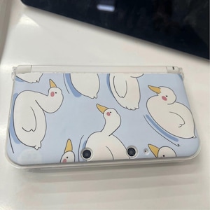 May include: A light blue handheld gaming console case featuring a pattern of white cartoon ducks with yellow beaks and pink cheeks. The ducks are set against a light blue background. The case has a clear plastic cover.